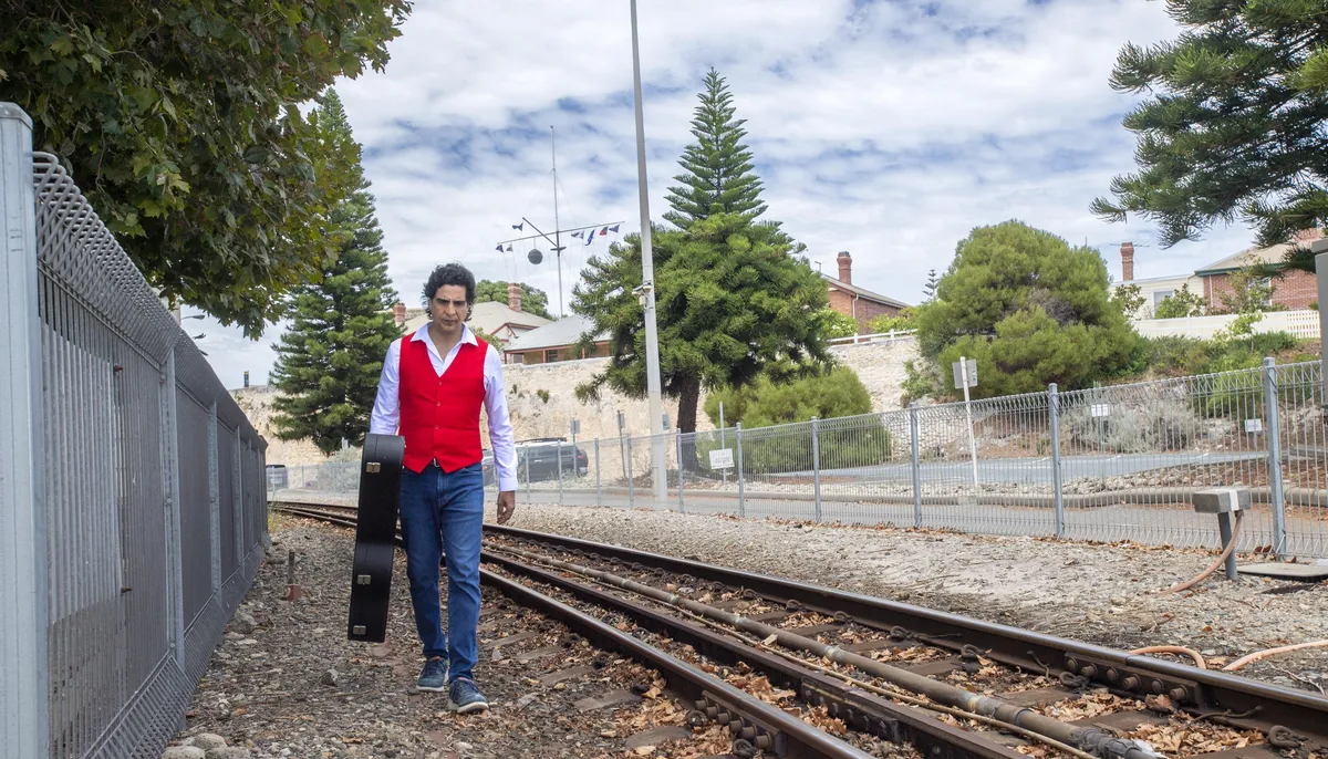 Ernesto Castillo in a red vest walking along a railway with his guitar case