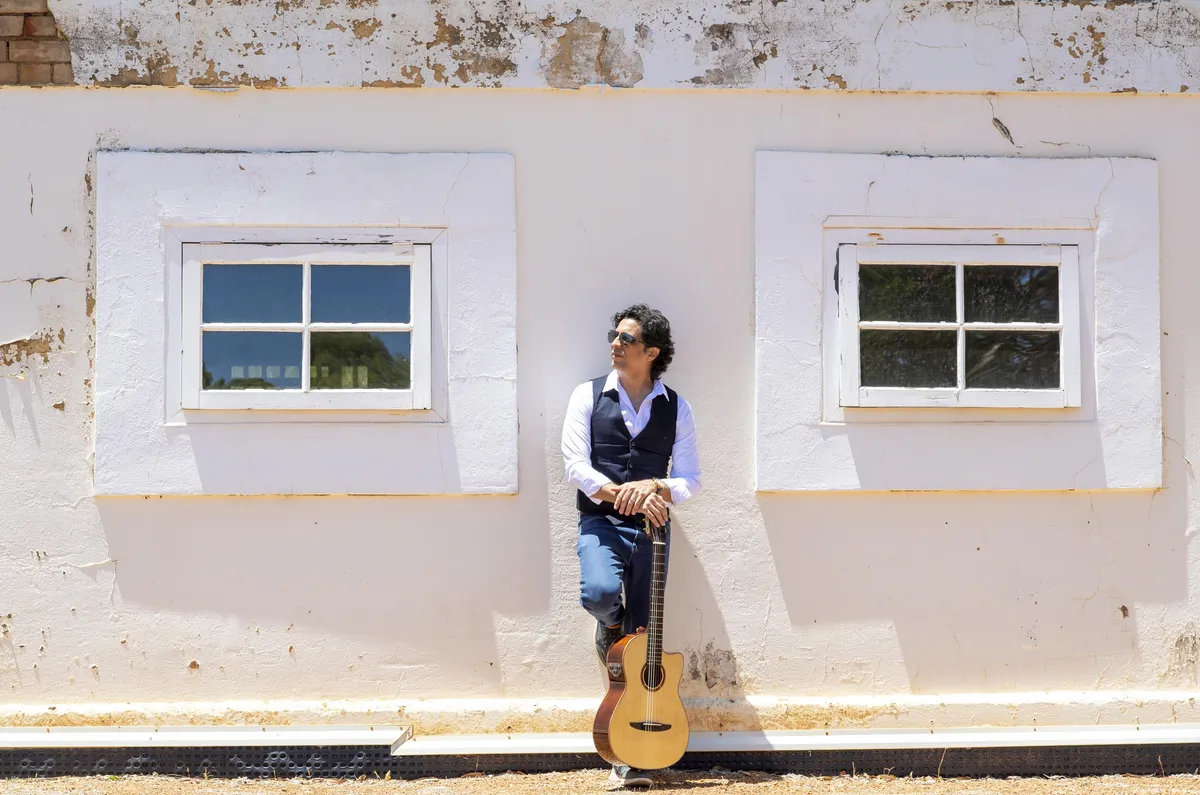 Ernesto Castillo in sunglasses leaning on a white wall with his guitar