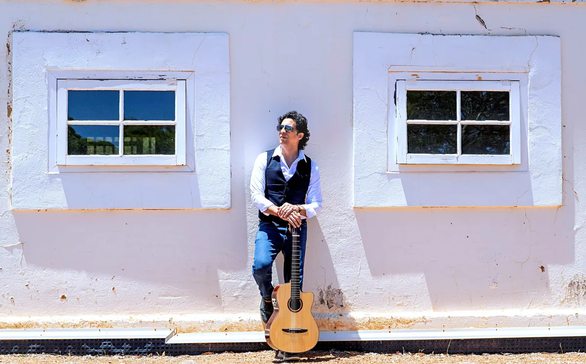 Ernesto Castillo leaning on a white wall between two windows with his guitar